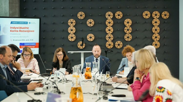 Grupa osób siedzących wokół stołu konferencyjnego w nowoczesnym biurze, zaangażowanych w spotkanie. Na ekranie prezentacji wyświetlane są informacje o webinarze inwestycyjnym.