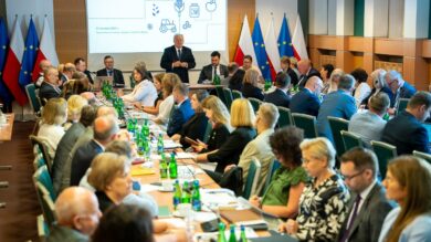 Duża grupa osób w strojach biznesowych siedzących wokół stołów konferencyjnych podczas spotkania, z mówcą stojącym z przodu i ekranem prezentacji wyświetlającym tekst za nim.