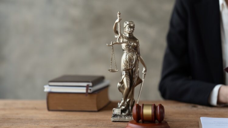 Pomnik Lady Justice, młotek, książki i część ramienia osoby w garniturze widoczne na drewnianym biurku.