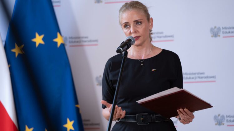 Kobieta ubrana na czarno, trzymająca czerwony segregator i mówiąca do mikrofonu, stoi przed flagami UE i Polski.