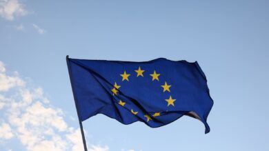Niebieska flaga z dwunastoma żółtymi gwiazdami w okręgu, reprezentująca Unię Europejską, powiewa na tle czystego nieba z kilkoma chmurami.