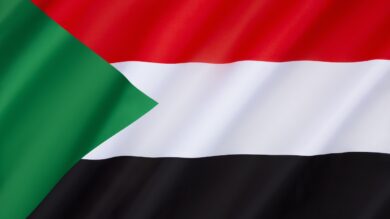 Zdjęcie przedstawia flagę Sudanu z poziomymi paskami w kolorze czerwonym, białym i czarnym oraz zielonym trójkątem po lewej stronie.