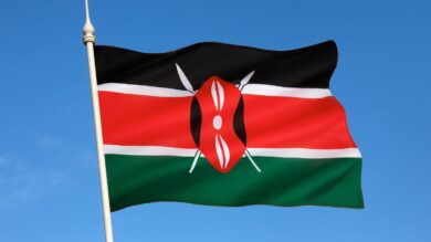 Flaga narodowa Kenii z czarnymi, czerwonymi i zielonymi paskami z białą obwódką oraz emblematem tarczy Masajów i włóczni powiewa na tle czystego, błękitnego nieba.