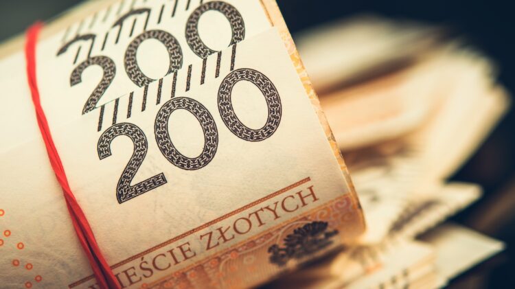 Stos zwiniętych banknotów 200 złotych polskich zabezpieczony czerwoną gumką. W tle widać więcej banknotów.
