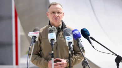 Mężczyzna w brązowej kurtce stoi na podium z wieloma mikrofonami z różnych serwisów informacyjnych. Wygląda na to, że przemawia na zewnątrz.