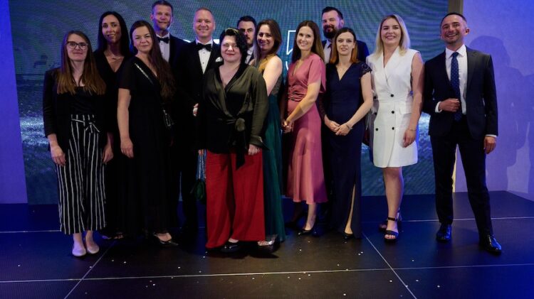 Grupa trzynastu osób, składających się z mężczyzn i kobiet, stoi razem na scenie ubrana w strój formalny i pozuje do zdjęcia.